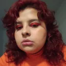 Mulher branca de cabelos vermelhos ondulados na altura dos ombros. Ela usa sombra vermelha e blusa laranja