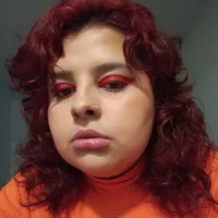 Mulher branca de cabelos vermelhos ondulados na altura dos ombros. Ela usa sombra vermelha e blusa laranja