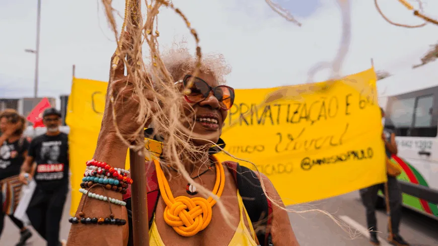 Mulher negra idosa, de óculos laranja, colar amarelo grosso e pulseiras coloridas sorri enquanto segura um mastro com fitilhos. Ao fundo, um grande pano amarelo com dizeres de protesto e pessoas marchando na 2ª Marcha das Mulheres Negras 2025, em Brasília.