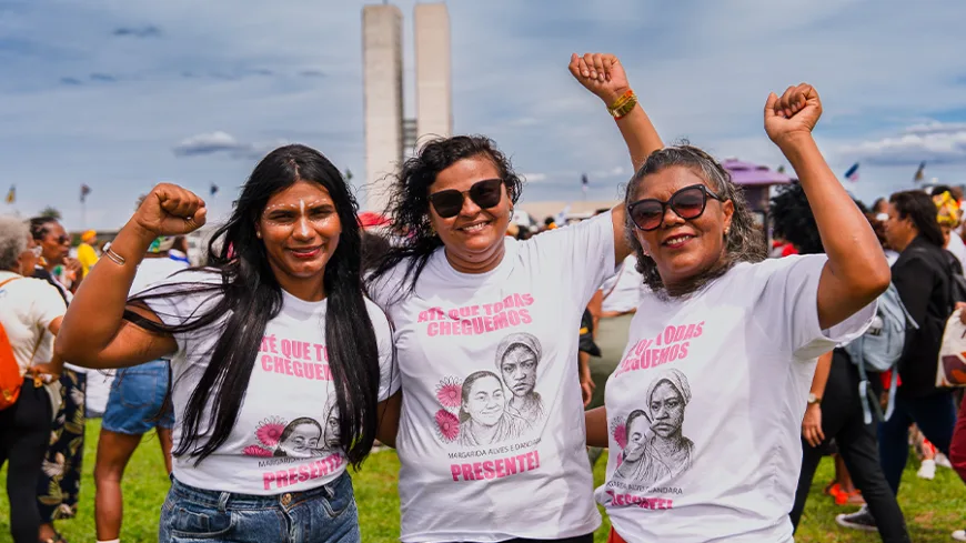 Três mulheres com camisetas brancas “ATÉ QUE TODAS CHEGUEMOS” posam juntas sorrindo. Todas elas estão com o punho levantado. Atrás delas, grande concentração de pessoas no gramado e as torres do Congresso ao fundo.