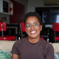 Beatriz de Oliveira é uma mulher negra, de cabelos escuros curtos, que usa óculos de grau. Na foto ela está sorrindo.