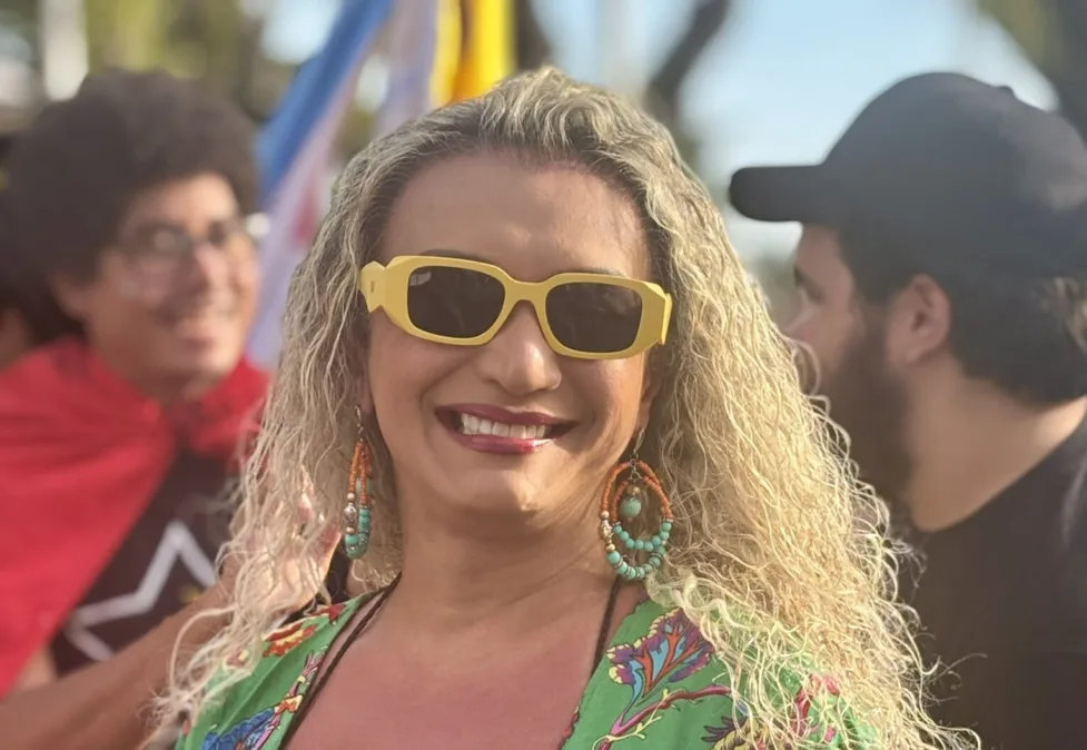 Retrato em plano fechado de uma pessoa sorrindo ao ar livre. Usa óculos de sol amarelos, cabelo loiro cacheado, brincos grandes com contas laranja e turquesa e blusa verde estampada. Ao fundo, desfocados, aparecem pessoas e bandeiras coloridas sob luz de fim de tarde.