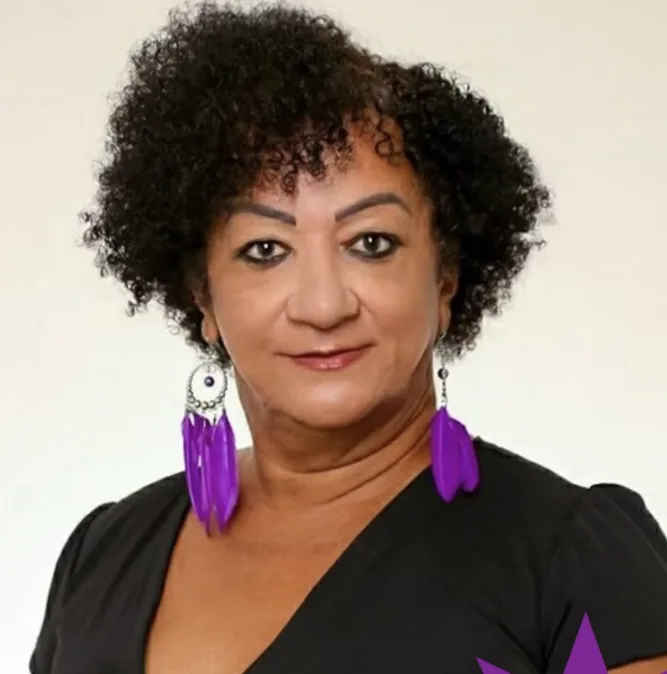 Retrato em plano médio de uma pessoa com cabelo curto e cacheado, sobrancelhas marcadas e maquiagem nos olhos. Veste blusa preta de decote em “V” e usa brincos longos roxos em formato de penas. Fundo neutro claro; expressão séria e confiante, olhando para a câmera.
