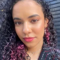 Uma mulher de cabelos castanhos escuros cacheados e longos, com pontas rosadas. Ela usa uma blusa preta