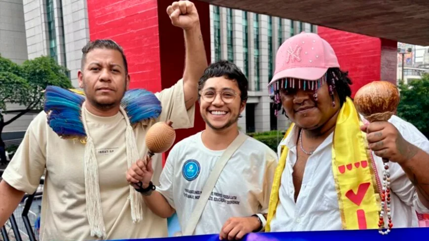 Três pessoas sorrindo e posando em frente ao MASP, na Av. Paulista. À esquerda, um homem com adorno de penas azuis sobre os ombros levanta o punho em gesto de luta; ao centro, pessoa de óculos com um maracá na mão; à direita, pessoa com boné rosa com franjas coloridas, lenço amarelo e cabaça/maracá nas mãos. Ao fundo, o vão livre vermelho do museu e prédios.