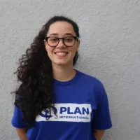 Mulher branca de cabelos cacheados escuros e longos. Ela usa óculos de grau e uma blusa azul da Plan International.