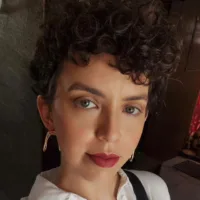 Mulher branca de cabelos castanhos escuros, cacheados e curtos. Usa camisa branca e brincos dourados.