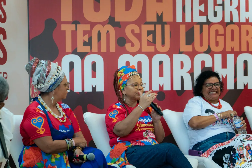 Três mulheres negras sentadas em um palco, participando de uma conversa pública. A mulher ao centro fala ao microfone. Ao fundo, um painel vermelho com textos grandes em português.