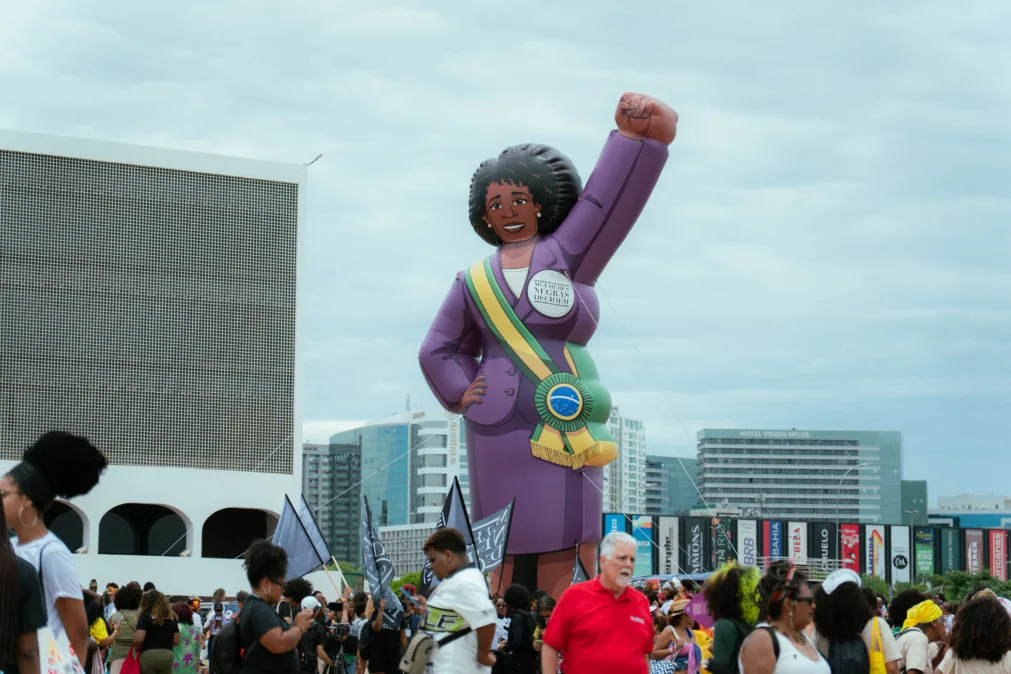 Boneca inflável gigante de uma mulher negra com punho erguido, vestindo terno roxo e faixa presidencial com a frase “Mulheres Negras Decidem”. Ao redor, multidão em manifestação, com bandeiras e prédios ao fundo.