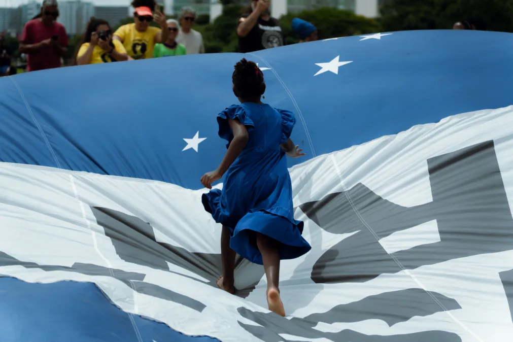 Criança negra, de vestido azul, corre descalça sobre uma grande bandeira inflável azul e branca com estrelas. Ao fundo, pessoas observam e fotografam a cena em um espaço aberto.