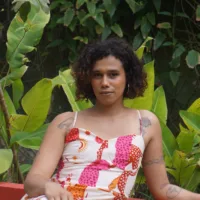 É uma mulher trans negra de pele clara, com cabelos cacheados escuros na altura do pescoço