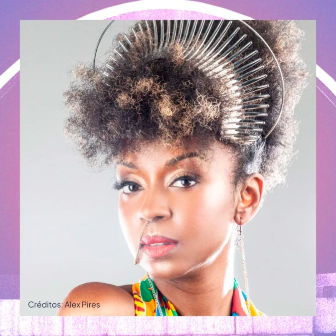 Retrato de uma mulher negra em close, olhando diretamente para a câmera. Ela tem cabelo crespo preso no alto da cabeça, com um adorno metálico em forma de arco e dentes que lembra um pente. Usa maquiagem suave, batom em tom rosado e um piercing delicado que liga o nariz ao lábio por uma corrente fina. Veste roupa colorida com estampa vibrante. O fundo é claro, emoldurado por tons de roxo e rosa. No canto inferior esquerdo, lê-se “Créditos: Alex Pires”.
