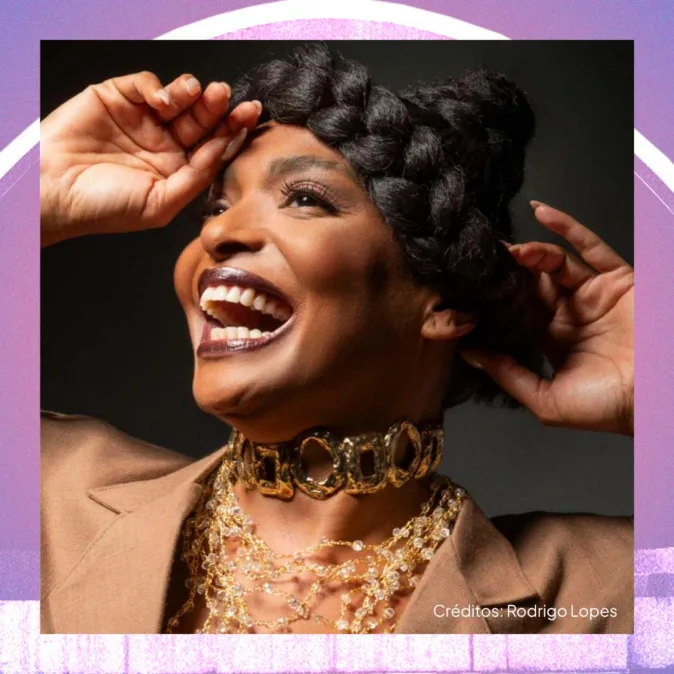 Retrato de uma mulher negra sorrindo amplamente, com a boca aberta e expressão de alegria. Ela olha levemente para cima, com as mãos erguidas próximas à cabeça. Usa cabelo trançado preso no alto, maquiagem marcante e batom escuro. Veste um blazer em tom neutro e vários colares dourados sobrepostos, incluindo uma gargantilha larga metálica. O fundo é escuro, criando contraste com a iluminação do rosto. A imagem é emoldurada por tons de roxo e rosa. No canto inferior direito, lê-se “Créditos: Rodrigo Lopes