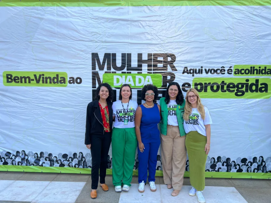 Cinco mulheres estão lado a lado, em pé, sorrindo para a câmera, diante de um grande painel branco com detalhes em verde. O painel traz os textos: “Bem-vinda ao”, “MULHER”, “DIA DA”, e, à direita, “Aqui você é acolhida e protegida”, com algumas palavras destacadas em verde. Na parte inferior, há uma ilustração com várias mulheres em preto e branco, formando uma faixa decorativa. Da esquerda para a direita: • A primeira mulher tem pele clara a média, cabelo escuro cacheado na altura dos ombros, e veste uma blusa vermelha com estampa floral e um casaco preto. • A segunda mulher tem pele clara, cabelo liso preso, e usa camiseta branca com estampa do evento e calça verde. • A terceira mulher é negra, com cabelo crespo curto e volumoso, usa óculos de armação clara, blusa azul sem mangas e calça azul, além de colar e brincos. • A quarta mulher tem pele clara, cabelo escuro longo e liso, usa óculos, camiseta branca do evento, colete verde e calça bege. • A quinta mulher tem pele clara, cabelo loiro longo e ondulado, usa óculos, camiseta branca do evento e calça verde. Todas estão em um espaço externo, sobre piso de pedra, com iluminação natural. A imagem transmite um ambiente institucional e acolhedor, relacionado a ações de apoio e proteção às mulheres.