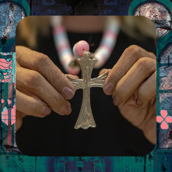 A imagem mostra um close das mãos de uma pessoa segurando um crucifixo metálico prateado. O objeto está centralizado e é sustentado delicadamente pelos dedos de ambas as mãos, com unhas curtas e naturais.

O crucifixo tem formato clássico, com detalhes em relevo, e no topo há uma conta esférica rosa que se destaca. Ao fundo, desfocado, é possível ver parte de um colar composto por contas grandes em tons de rosa, branco e azul — cores associadas à bandeira trans.

A pessoa veste uma roupa preta, que serve de fundo neutro para o objeto. A iluminação é suave e quente, destacando o brilho metálico do crucifixo e a textura das mãos.

A imagem está inserida em uma moldura gráfica com cantos arredondados, em tons de azul e verde, com texturas que lembram vitrais ou colagens, reforçando uma estética simbólica que dialoga com religiosidade e identidade