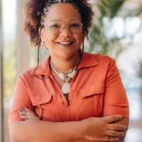 mulher negra com cabelos castanhos crespos e curtos, vestido toto laranja, de oculos e pracos cruzados, sorrindo