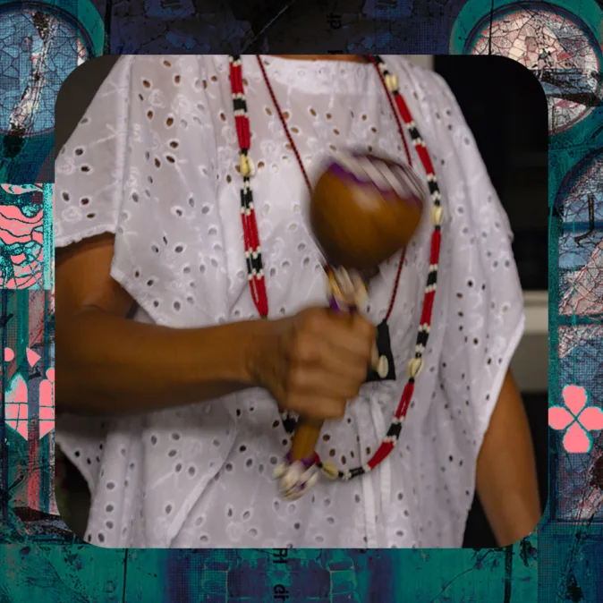 Imagem em close mostra o tronco de uma pessoa, com foco nas mãos e no objeto que segura. A pessoa veste uma roupa branca rendada, com textura vazada e delicada, semelhante a vestimentas tradicionais de matriz religiosa afro-brasileira. No pescoço, usa colares longos de contas nas cores vermelho, preto e branco, que descem sobre o peito.

Na mão direita, segura um instrumento de madeira arredondado, semelhante a um maracá ou chocalho, em movimento — o que cria um leve desfoque na imagem, indicando que está sendo sacudido.

O fundo da imagem é escuro e desfocado, destacando o corpo, a roupa e o objeto ritual. A cena é enquadrada dentro de uma moldura gráfica com elementos visuais em tons de azul e verde, com texturas e padrões abstratos que lembram vitrais ou grafismos.

A imagem transmite a ideia de prática espiritual ou ritual, com ênfase nos elementos simbólicos e no gesto em movimento.