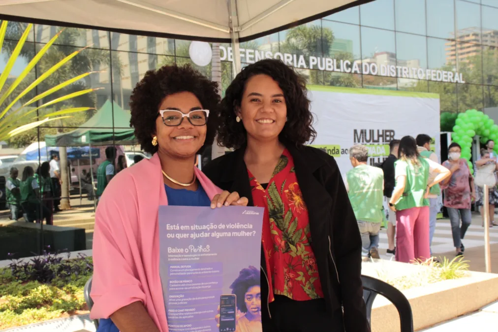 Duas mulheres estão lado a lado, em primeiro plano, sorrindo para a câmera sob uma tenda branca em um evento ao ar livre. À esquerda, uma mulher negra, de pele média-escura, com cabelo crespo volumoso em formato arredondado. Ela usa óculos de armação clara, brincos dourados e um colar, além de uma blusa azul com um casaco rosa por cima. Sua expressão é acolhedora e confiante. À direita, uma mulher de pele clara a média, com cabelo escuro, cacheado e na altura dos ombros. Ela veste uma blusa vermelha com estampa floral e um casaco preto. Também sorri diretamente para a câmera. As duas seguram juntas um cartaz em tons de roxo e azul. O texto principal diz: “Está em situação de violência ou quer ajudar alguma mulher? Baixe o PenhaS”. O cartaz apresenta funcionalidades do aplicativo, como manual de fuga, botão de pânico, gravação de áudio, rede de apoio e pontos de apoio, além da imagem de um celular com a interface do app. Ao fundo, há um espaço movimentado com outras pessoas, algumas vestindo coletes verdes, e uma faixa com o texto “Defensoria Pública do Distrito Federal”. Também aparecem elementos urbanos, como prédios, árvores e uma área com plantas, indicando um evento institucional voltado ao atendimento e orientação da população.