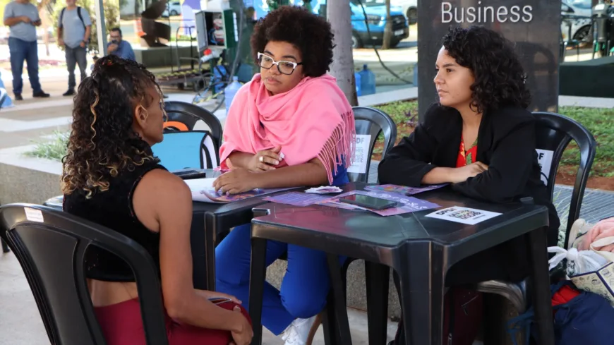 Três mulheres estão sentadas ao redor de uma mesa preta, em um ambiente externo, participando de uma conversa. Em primeiro plano, de costas para a câmera, está uma mulher de pele média, com cabelo cacheado preso em um rabo de cavalo. Ela usa uma blusa preta sem mangas e calça vermelha, e está voltada para as outras duas mulheres, em posição de escuta e diálogo. À frente dela, do outro lado da mesa, estão duas mulheres. À esquerda, uma mulher negra, de pele média-escura, com cabelo crespo volumoso e óculos de armação clara. Ela usa um xale rosa sobre os ombros, calça azul e tênis branco. Sua expressão é atenta e acolhedora, com o corpo levemente inclinado para a frente, indicando escuta ativa. À direita, uma mulher de pele clara a média, com cabelo escuro cacheado na altura dos ombros. Ela veste uma blusa vermelha com estampa floral e um casaco preto. Está com os braços apoiados na mesa, também voltada para a mulher à sua frente, com expressão concentrada. Sobre a mesa, há materiais informativos, folhetos em tons de roxo, um celular e um pequeno cartaz com QR code, sugerindo orientação ou atendimento. Ao fundo, há um espaço público com circulação de pessoas, árvores e veículos. Um painel vertical exibe o texto “Esplanada Business”. A cena transmite um contexto de atendimento ou acolhimento em um evento ao ar livre, possivelmente voltado à orientação e apoio a mulheres.