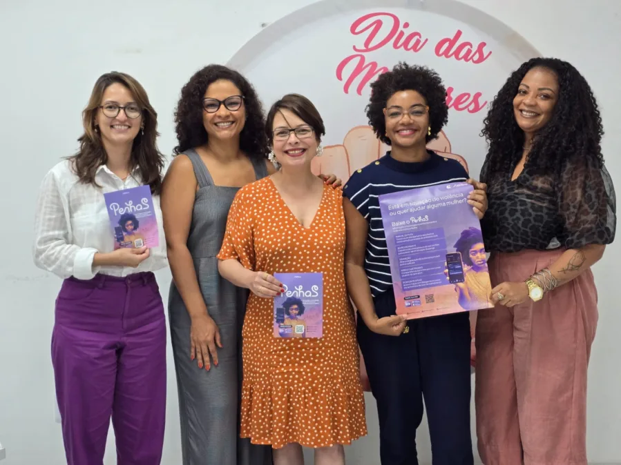 Cinco mulheres estão lado a lado, em pé, sorrindo para a câmera, em um ambiente interno com parede clara. Ao fundo, há um painel circular com o texto “Dia das Mulheres” em letras rosa. Da esquerda para a direita: • A primeira mulher tem pele clara, cabelo castanho claro na altura dos ombros e usa óculos. Veste uma camisa branca e calça roxa, e segura um folheto do aplicativo PenhaS. • A segunda mulher tem pele média, cabelo escuro cacheado e usa óculos. Veste um vestido cinza sem mangas. • A terceira mulher tem pele clara, cabelo curto e liso, usa óculos e veste um vestido laranja com estampa de bolinhas brancas. Ela também segura um folheto do PenhaS. • A quarta mulher é negra, com cabelo crespo curto e volumoso, usa óculos e veste uma blusa azul escura com detalhes claros. Ela segura um cartaz maior do PenhaS com informações sobre o aplicativo. • A quinta mulher tem pele média, cabelo longo e cacheado, e veste uma blusa preta transparente com estampa e uma calça em tom rosado. Os materiais exibidos destacam o aplicativo PenhaS, voltado ao apoio de mulheres em situação de violência, com textos e imagens em tons de roxo e rosa. A cena transmite um ambiente institucional e de mobilização em torno do Dia das Mulheres.