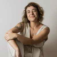 Maria é branca, tem cabelos loiros e está sentada com o joelho dobrando vestindo uma roupa clara.