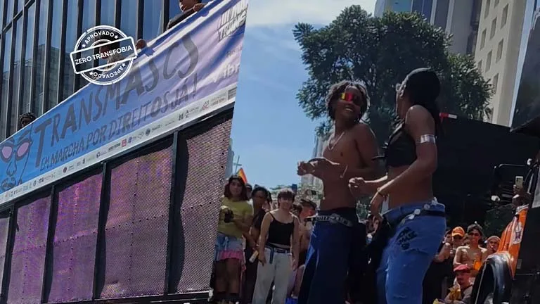A imagem está dividida em duas partes, mostrando momentos de uma marcha em espaço urbano, sob luz do dia. À esquerda, aparece um caminhão de som com um grande banner estendido na lateral. O banner traz a palavra “TRANSMASCULINIDADES” em destaque e a frase “em marcha por direitos”, além de logotipos de organizações. Sobre a imagem, há um selo gráfico com a frase “Zero Transfobia”. Ao fundo, prédios altos sugerem que a cena acontece em uma área central da cidade. À direita, duas pessoas dançam sobre o caminhão ou em uma estrutura elevada. Uma delas está sem camisa, usando óculos escuros coloridos e calça larga; a outra veste um top preto e calça jeans. Ambas parecem jovens adultas, com corpos em movimento e expressões de alegria e energia.