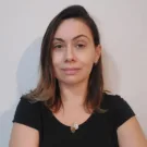 Priscila é branca, tem cabelos castanhos claros e lisos um pouco abaixo dos ombros, e está usando uma blusa preta