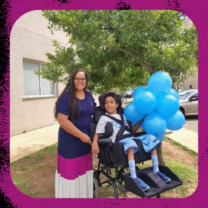 Eliana tem cabelos longos ondulados, usa óculos e está acompanhada do filho, que está numa cadeira de rodas.