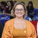 Gisa é uma mulher branca, de cabelos loiros, que usa óculos