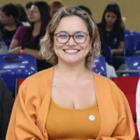 Gisa é uma mulher branca, de cabelos loiros, que usa óculos
