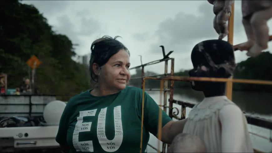 Frame do documentário Guardiãs do Futuro. A imagem apresenta tons de verde, cinza e azul, sob luz natural e céu nublado. Em primeiro plano, uma mulher de meia-idade, de pele morena e cabelos escuros presos em um coque, está em uma embarcação às margens de um rio. Ela veste uma camiseta verde com grandes letras brancas formando a palavra “EU”, preenchidas por palavras menores. A mulher apoia um dos braços na estrutura da embarcação e olha para a direita da imagem com expressão contemplativa. Ao seu lado há uma instalação artística composta por bonecos e manequins. Um deles aparece em destaque: uma figura infantil de rosto escuro e vestido claro. Outros bonecos surgem parcialmente no canto superior direito. Ao fundo, vê-se o rio cercado por vegetação densa e árvores, além de estruturas metálicas enferrujadas da embarcação. O céu está coberto por nuvens cinzentas, reforçando a atmosfera ambiental e documental da cena.