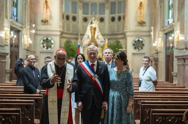 Fotografia mostra um grupo caminhando pelo corredor central de uma igreja católica ornamentada. Em destaque, um homem grisalho usando faixa presidencial vermelha, branca e azul caminha de mãos dadas com uma mulher de vestido azul-claro estampado. Ao lado deles, um cardeal com vestes pretas e vermelhas gesticula enquanto conversa. Outras pessoas acompanham o grupo ao fundo, entre bancos de madeira e altares iluminados. A cena transmite solenidade, religião e poder político institucional.