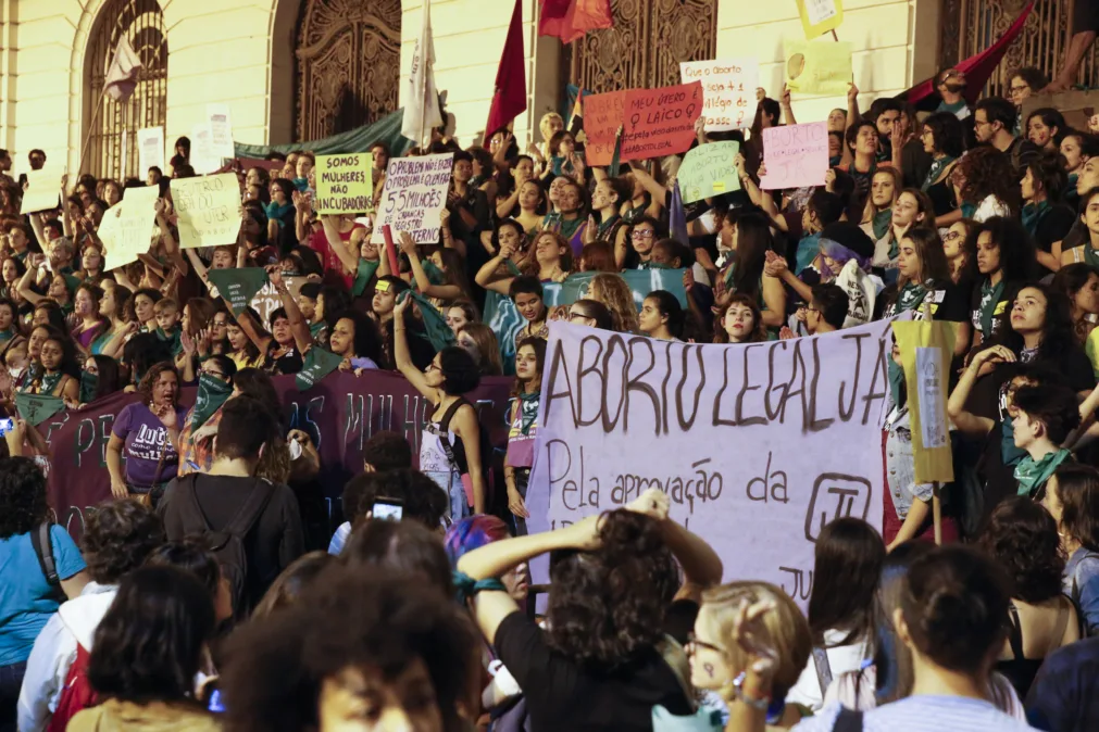 Fotografia mostra uma grande manifestação feminista em frente a um prédio histórico à noite. Centenas de pessoas ocupam escadarias e a rua segurando cartazes, faixas e bandeiras em defesa da legalização do aborto. Em destaque, uma faixa branca com letras grandes diz “ABORTO LEGAL JÁ”. Outros cartazes trazem frases como “Meu útero é laico” e “Aborto livre, legal e seguro”. Muitas participantes usam lenços verdes, símbolo da luta pelo direito ao aborto na América Latina. A cena transmite mobilização coletiva, protesto e reivindicação por direitos reprodutivos.