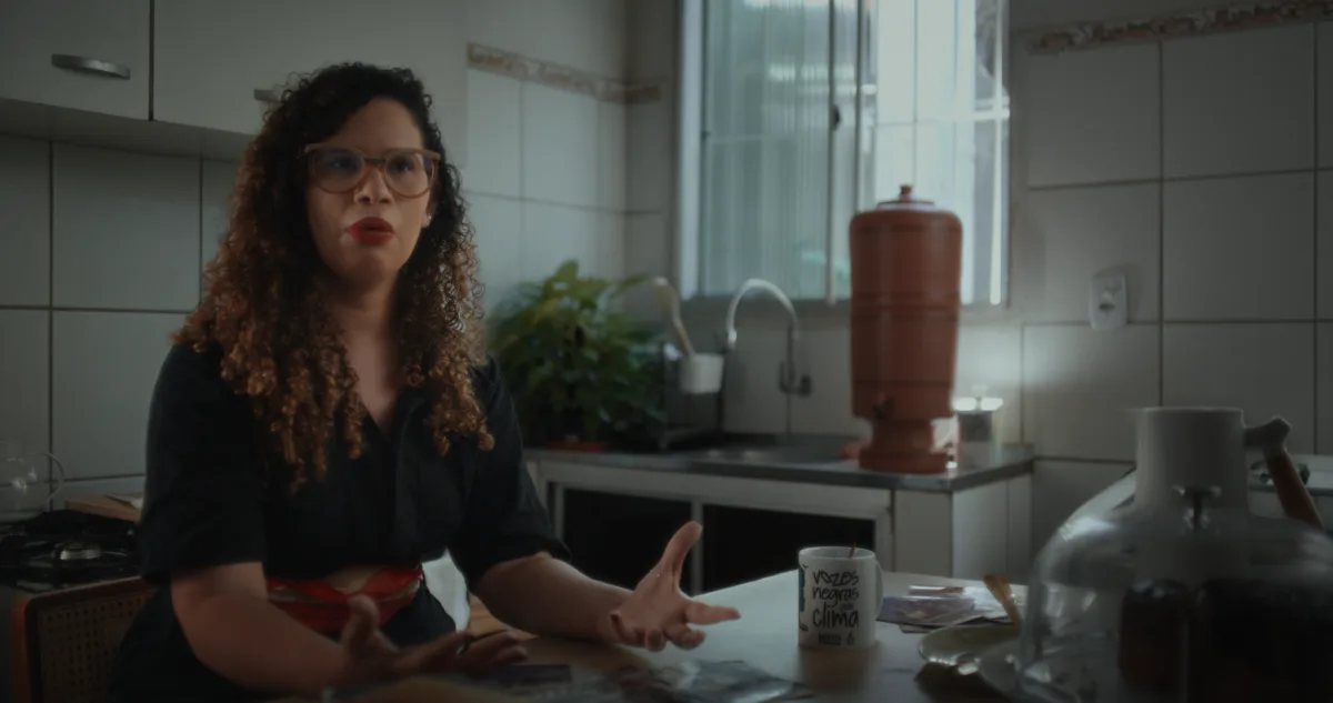 Frame do documentário Guardiãs do Futuro. Uma mulher de pele clara, cabelos cacheados longos e óculos de armação transparente está sentada à mesa de uma cozinha enquanto fala e gesticula com as mãos.

Ela veste uma camisa preta. Sobre a mesa há fotografias espalhadas, uma caneca branca com a frase “Vozes negras pelo clima”, pratos e recipientes de vidro. Ao fundo aparecem uma pia, uma janela iluminada por luz natural, uma planta verde e um filtro de barro. A cena tem tons suaves de cinza, bege e verde, com iluminação que entra pela janela e destaca o ambiente doméstico.