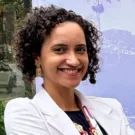 Natalia é uma mulher negra de pele clara, com cabelos cacheados curtos e castanho escuro