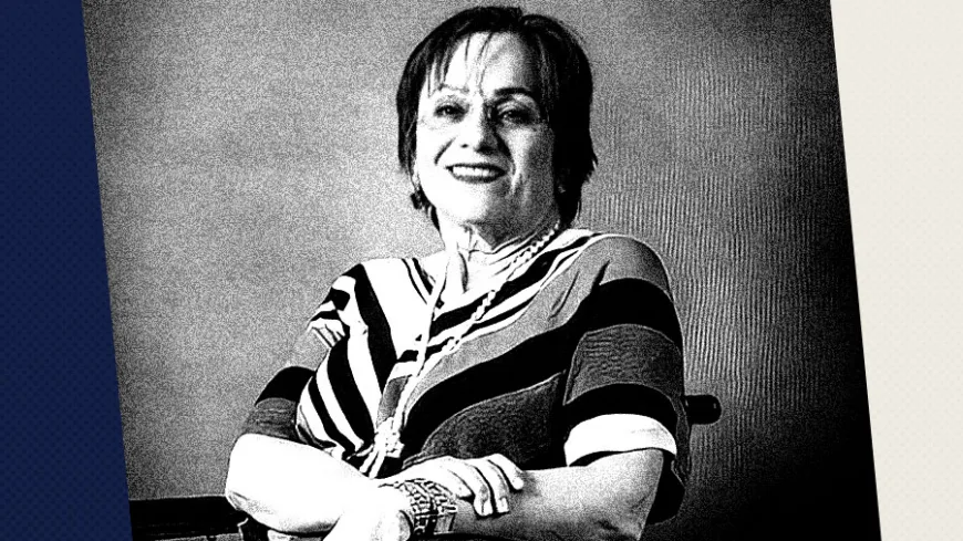 Fotografia em preto e branco de Maria da Penha, mulher de cabelos curtos sorrindo para a câmera. Ela está sentada, com os braços cruzados sobre o colo, usando uma blusa de mangas curtas com listras e formas geométricas, além de pulseiras e um colar longo. Ao fundo há uma parede lisa, enquanto faixas verticais em azul e branco enquadram a composição. O retrato transmite uma imagem de confiança e remete à atuação pública e política da personagem retratada.