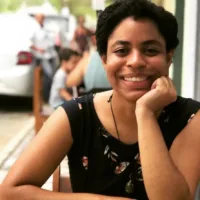 Mulher negra não retinta, de cabelos pretos, usando blusa preta. Ela sorri e está com a mão no queixo