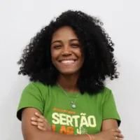 Taciere é uma mulher negra de cabelos pretos crespos, ela está sorrindo e usa uma blusa verde