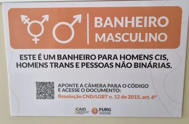 Fotografia de uma placa indicativa de "BANHEIRO MASCULINO", ilustrada com os símbolos masculino e transgénero. No centro, a mensagem principal destaca-se em letras grandes: "ESTE É UM BANHEIRO PARA HOMENS CIS, HOMENS TRANS E PESSOAS NÃO BINÁRIAS."