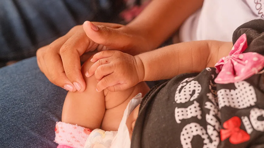 Fotografia em close de um bebê usando uma roupa preta com estampas brancas e vermelhas e um laço rosa na cabeça. O bebê está deitado, com as pernas parcialmente à mostra e uma das mãos apoiada sobre a própria perna. Ao lado, uma pessoa adulta segura delicadamente a perna do bebê com uma das mãos. A cena tem iluminação quente, com tons de pele alaranjados e fundo desfocado.