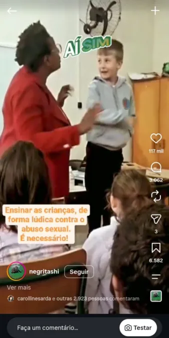 A imagem é uma captura de um vídeo em formato vertical de uma rede social. A cena decorre no interior de uma sala de aula. No centro da ação, está uma professora orientando um aluno a como impedir alguém de tocá-lo nas partes íntimas.