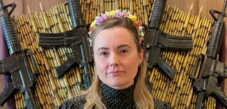 Uma mulher branca de cabelos loiros, a deputada Julia Zanatt (PL), usa uma tiara de flores coloridas e uma blusa preta de gola alta com bolinhas brancas e um colar de cruz prateada. Ela está posicionada à frente de um painel decorativo composto por dezenas de cartuchos de munição dispostos em arco e quatro fuzis pretos dispostos nas diagonais.